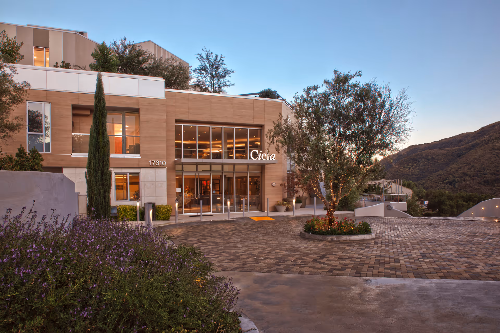 Front entrance of the Ciela senior living building with a paved driveway, lit glass doors, landscaping and hills in the background.