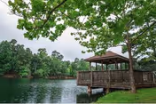 A wooden gazebo extends over a calm lake shore surrounded by trees and greenery.