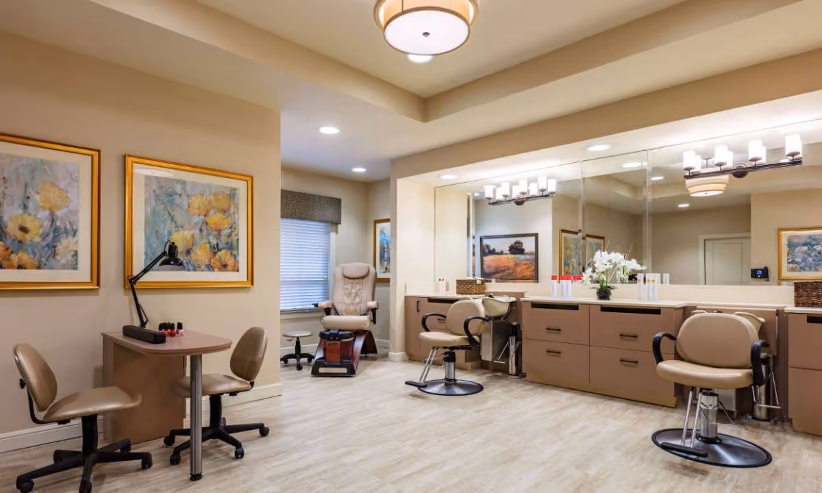 A well-lit salon area in a senior living facility featuring two salon chairs in front of a large mirror with overhead lighting, a pedicure chair near a window with blinds, and a small table with two chairs and a desk lamp. The walls are decorated with framed floral paintings.