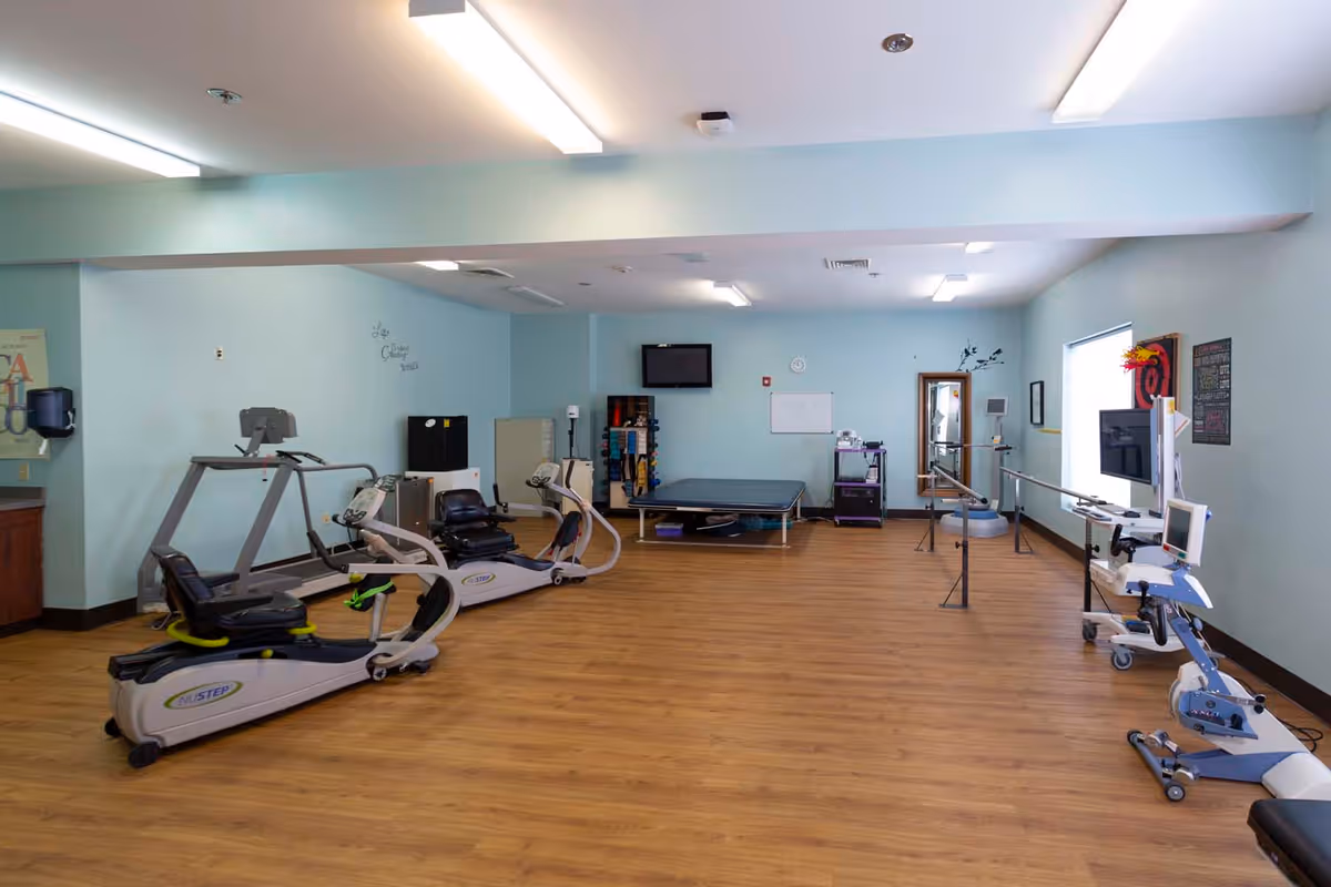 Therapy gym room with recumbent exercise machines, a treatment table, parallel bars and medical equipment in a rehabilitation facility.
