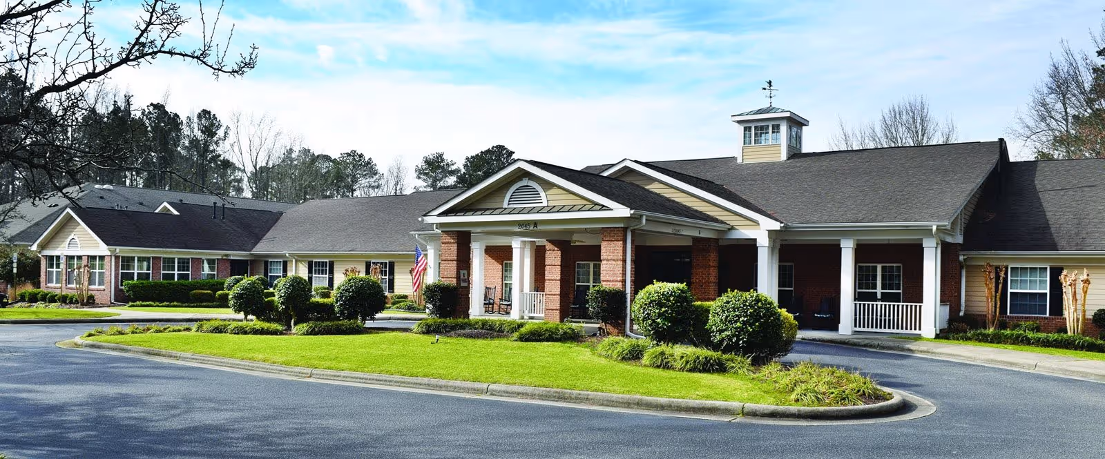 Front entrance of a single-story senior living facility with a covered portico, circular driveway, American flag, and manicured landscaping.