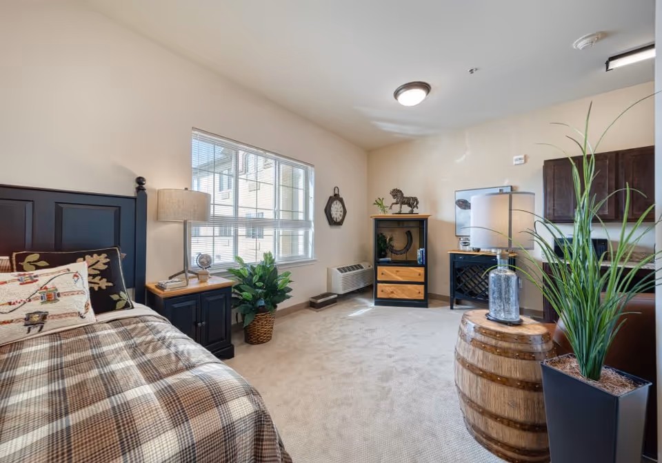 A cozy senior living bedroom with a plaid bedspread, decorative pillows, a bedside table with a lamp, a large window letting in natural light, a potted plant, a wall clock, a small cabinet with decorative items, and a wooden barrel used as a table with a lamp and a tall potted plant beside it. The room has beige walls and carpeted floor.