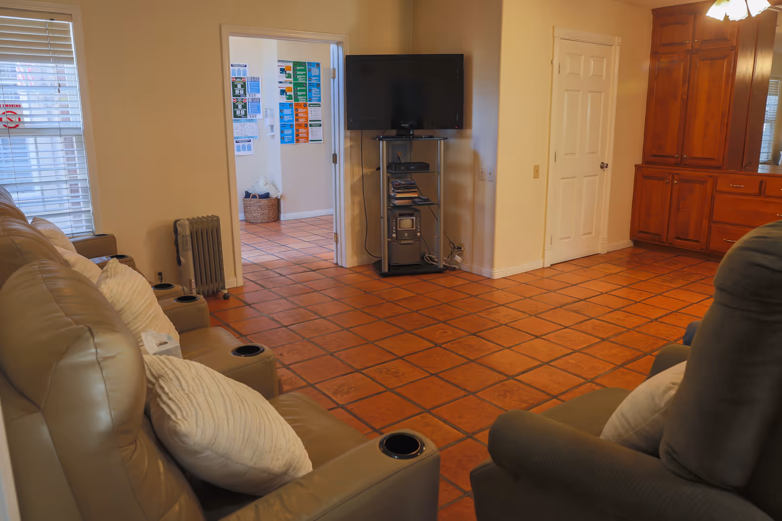 Interior living room with terracotta tile floor, recliner sofas and pillows, a wall-mounted TV on a stand, and wooden cabinets.
