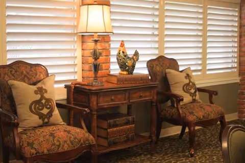Two ornate upholstered chairs flank a wooden side table with a lamp and decorative rooster figurine.