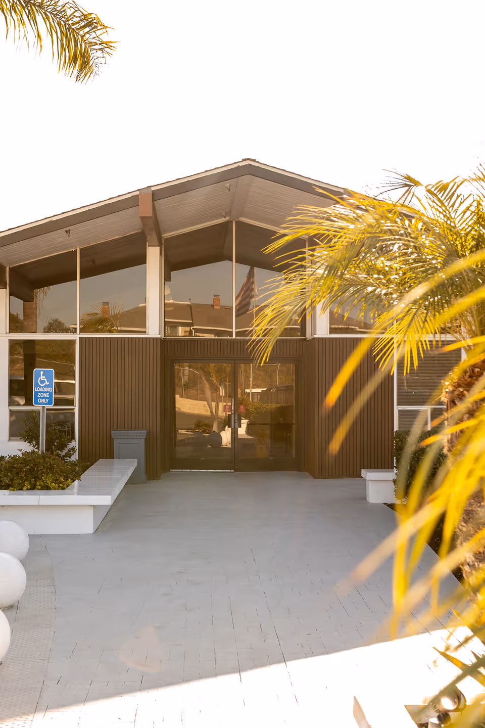 Exterior front entrance of a care facility with double glass doors, large upper windows, and palm trees flanking the walkway.