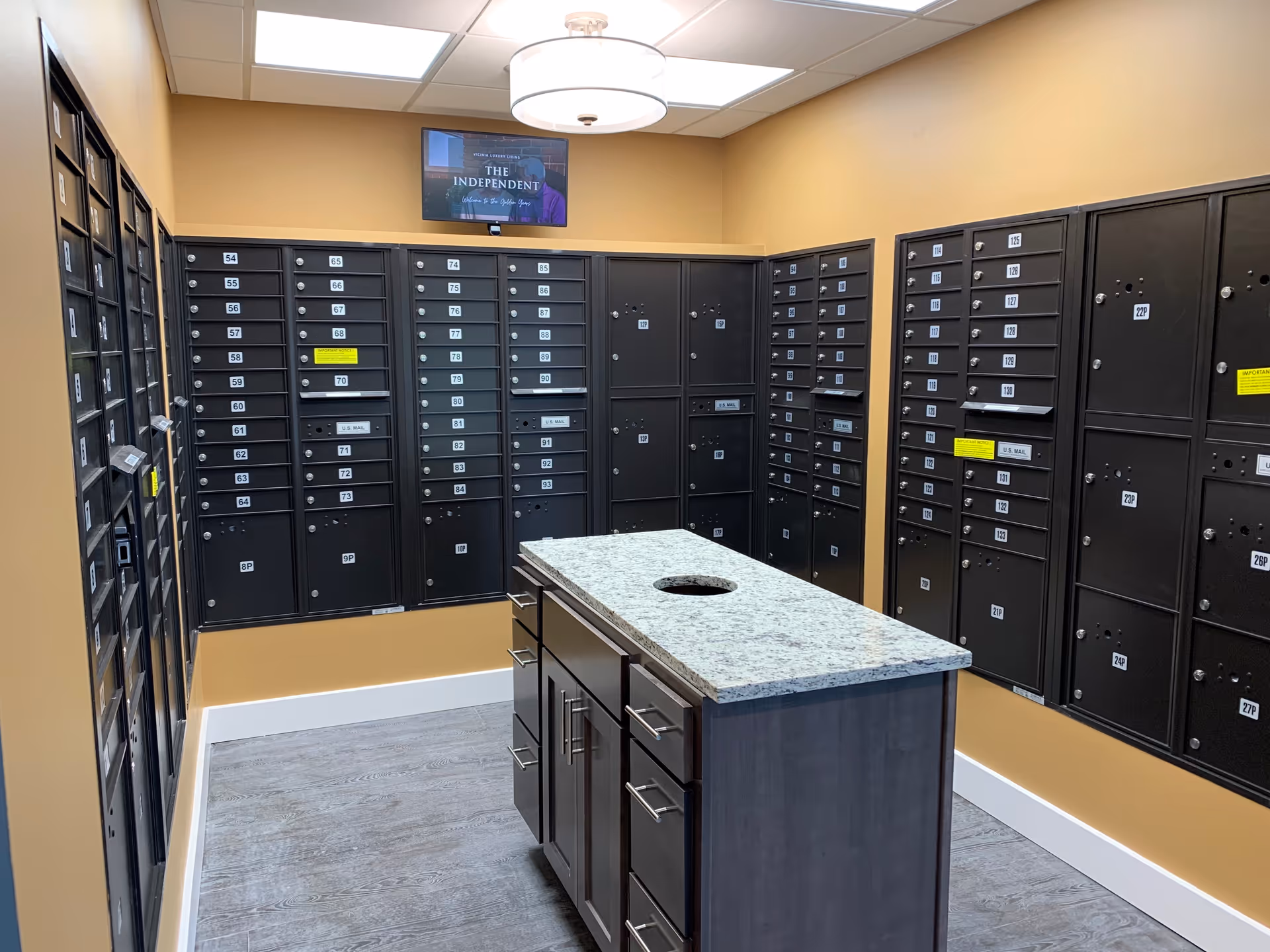 Interior view of a mailroom with multiple black mailboxes mounted on beige walls. There is a central island with a granite countertop and cabinets underneath. A ceiling light fixture and a TV screen displaying 'THE INDEPENDENT' are visible above the mailboxes.