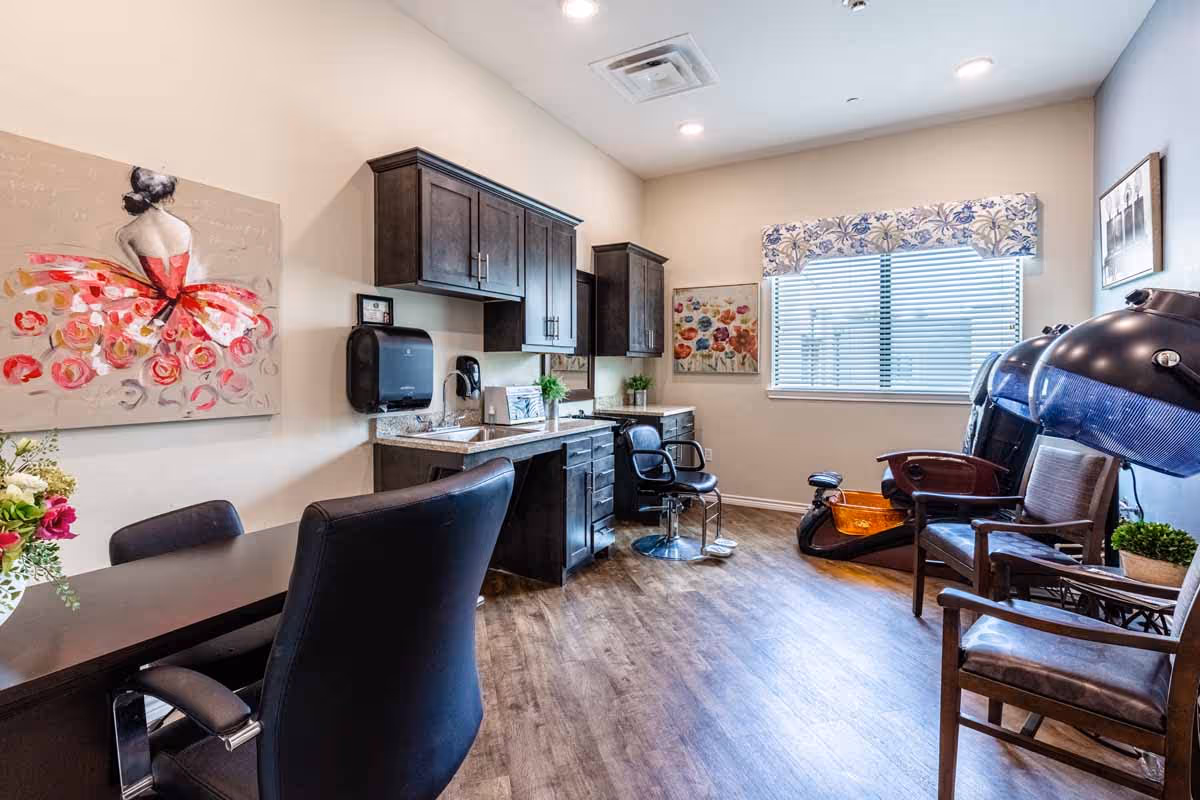 A bright salon room with wooden flooring, featuring a black salon chair in front of a counter with a sink and dark wooden cabinets above and below. There are two hair drying stations with hooded dryers and chairs on the right side near a window with blinds and a floral valance. The room is decorated with colorful paintings and a vase of flowers on a table with black chairs.
