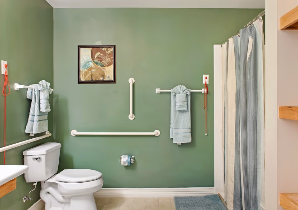 A bathroom with green walls featuring a white toilet, grab bars, a towel rack with folded blue towels, a shower with a blue and white striped curtain, a blue bath mat, and a framed artwork on the wall.