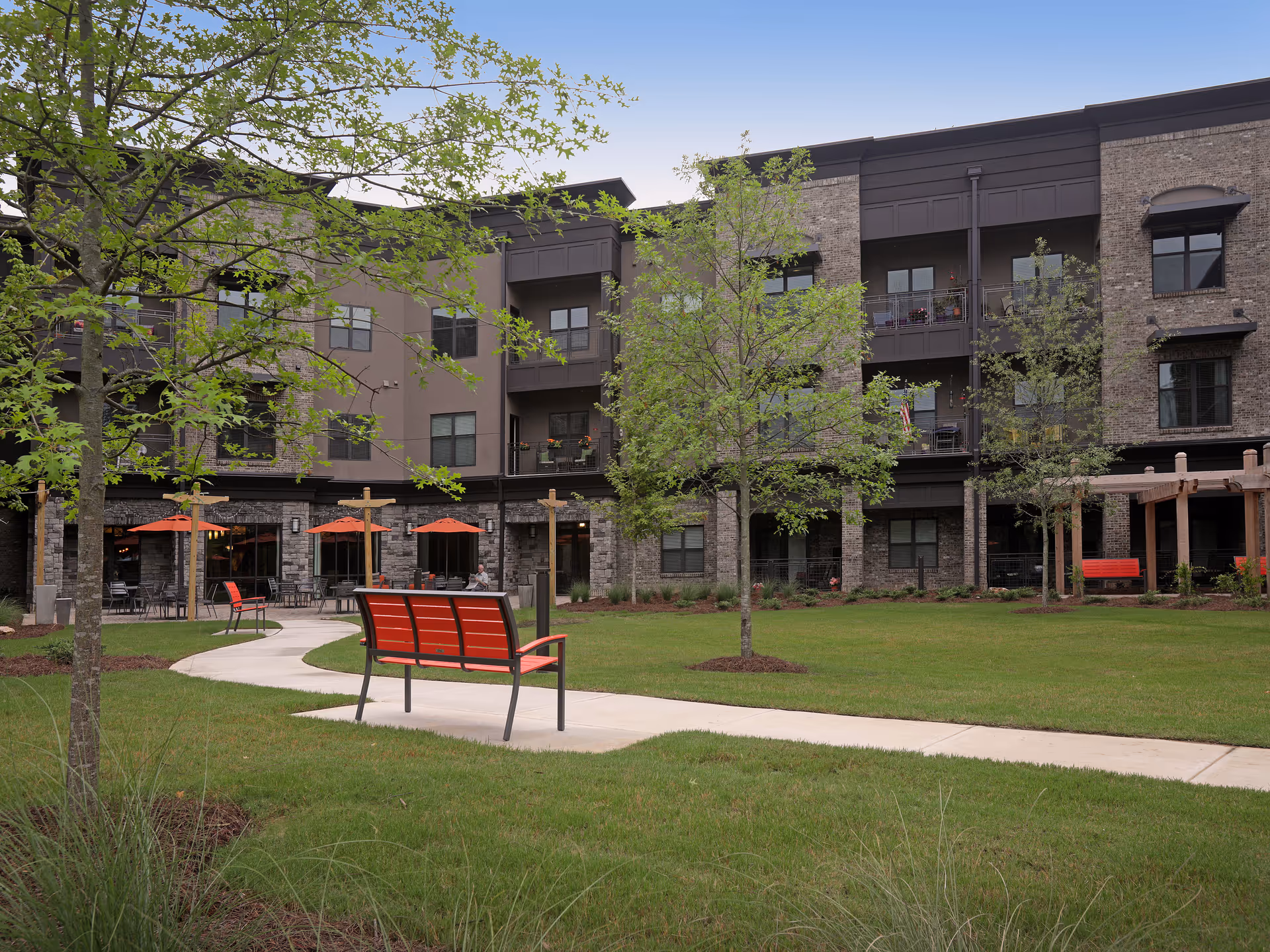 Outdoor courtyard of a multi-story senior living building with benches, a paved walkway, trees, and patio umbrellas.