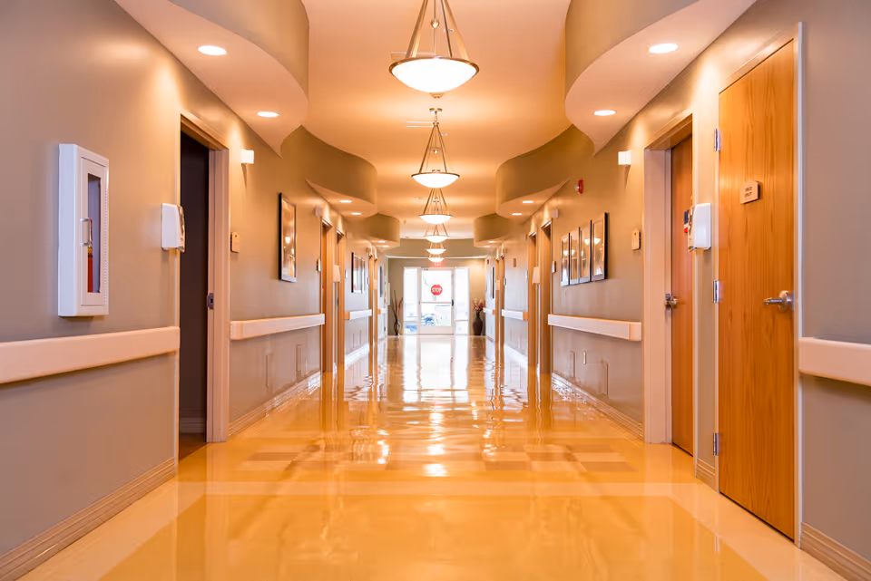 A long, clean hallway in a healthcare facility with polished yellow floors, beige walls, wooden doors on both sides, ceiling lights, and handrails along the walls.