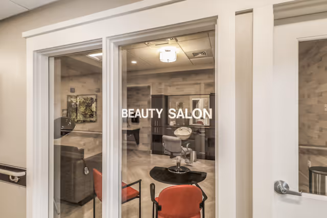 Interior view through glass doors labeled 'BEAUTY SALON' showing a beauty salon room with a salon chair, a black circular rug, cabinets, and a mirror. There are also two red chairs visible in the foreground outside the salon.