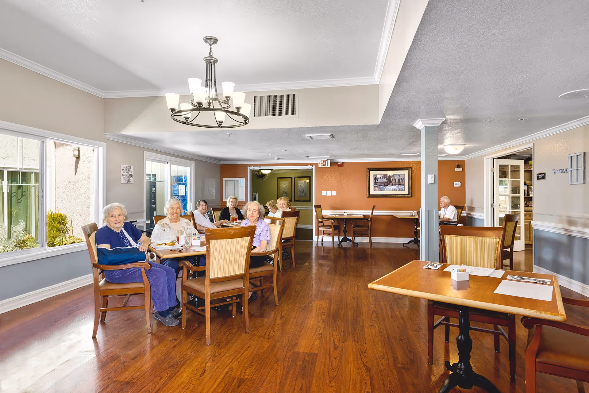 Elderly residents seated around tables in a bright assisted-living dining/activity room.