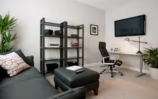 A modern office or study room with a black sofa and ottoman on the left, a black metal bookshelf with books and decorative items against the wall, a white desk with a black office chair, a desk lamp, and a flat-screen TV mounted on the wall. There are also two potted plants and framed pictures on the walls.