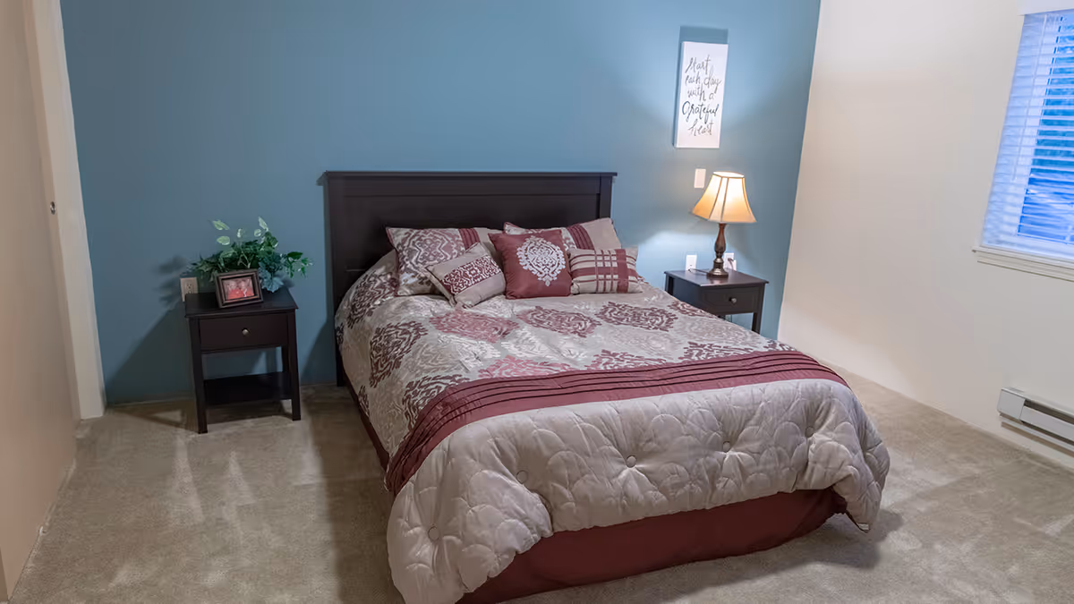 A neatly made bed with patterned bedding and multiple pillows in a bedroom. The bed has a dark wooden headboard and is flanked by two matching nightstands. One nightstand has a small plant and a framed photo, while the other has a lamp and a small decorative item. The walls are painted in two tones, with one wall teal and the others off-white. A window with blinds is visible on the right side of the room.