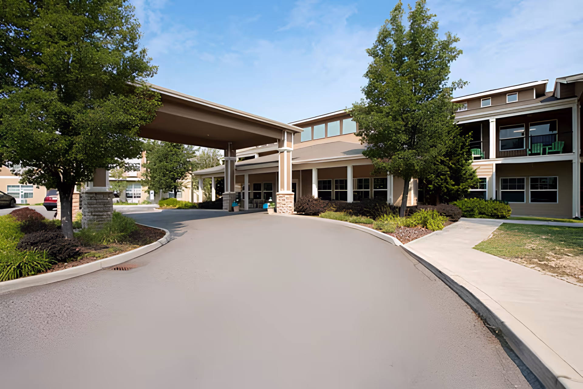 Front exterior view of Wenatchee Senior Living by Cogir showing the entrance driveway with a covered drop-off area, surrounded by trees and landscaping under a clear blue sky.