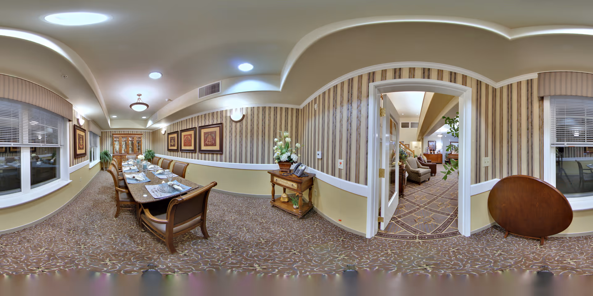 Long formal dining room with a set table, upholstered chairs, patterned carpet, and an adjacent sitting area visible through an open doorway.