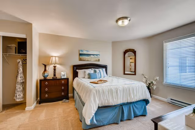 A neatly made bed with white and blue bedding in a bedroom. There is a wooden nightstand with a lamp and a framed photo next to the bed. A painting hangs above the headboard. A mirror is mounted on the wall near a window with blinds. An open closet with hanging clothes is visible to the left.