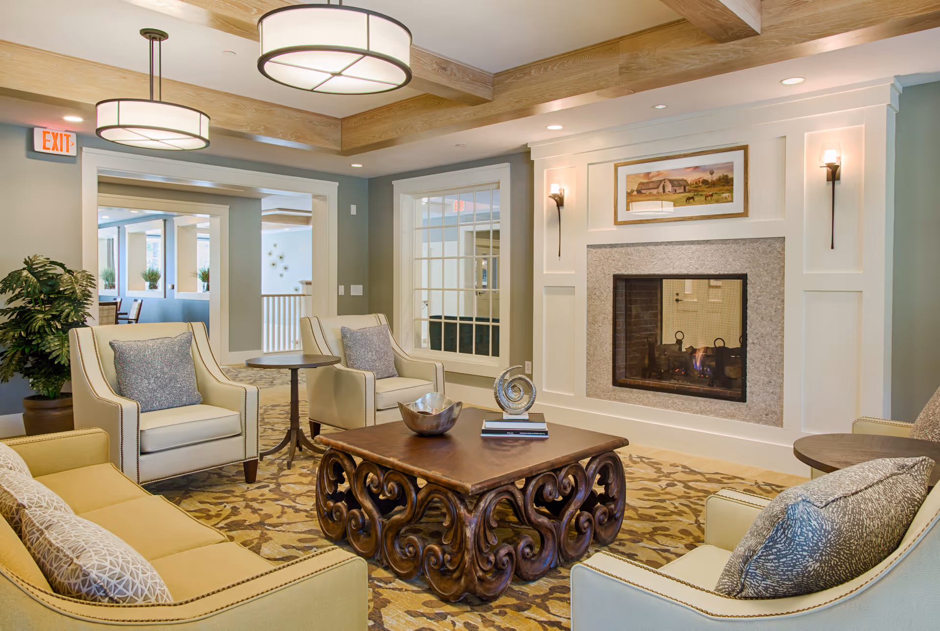 A cozy living room area in a senior living facility featuring a fireplace with a framed painting above it, two beige armchairs with gray cushions, a yellow sofa with patterned pillows, and a decorative wooden coffee table with a bowl and sculpture on top. The room has a patterned carpet, wooden ceiling beams, and modern round ceiling lights. There is a large window and an exit sign visible in the background.