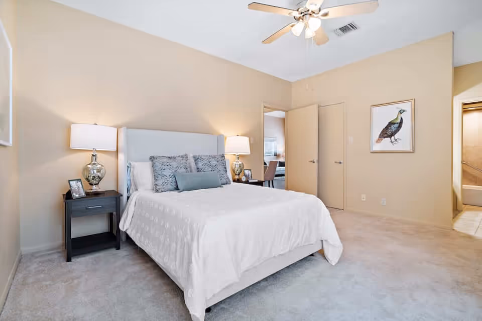 A bedroom with a neatly made bed featuring white bedding and multiple pillows. There are two bedside tables with matching lamps on either side of the bed. The walls are painted beige, and there is a ceiling fan with lights above. A framed picture of a bird hangs on the wall to the right, and two doors are visible, one leading to another room and the other to a bathroom.