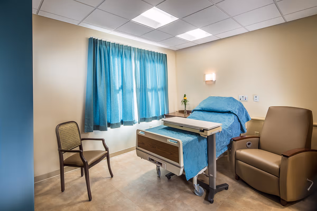 A small, clean room with a hospital-style bed covered in a blue blanket, a brown armchair, a wooden chair with a cushioned seat, a small side table with a flower vase, and a window with blue curtains.