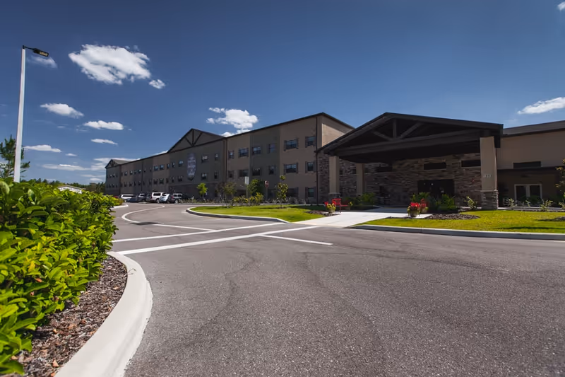 Exterior view of a large senior living facility building under a clear blue sky with a few clouds. The building has three stories with multiple windows and a covered entrance supported by stone pillars. There is a curved driveway leading to the entrance, landscaped with green bushes and grass.