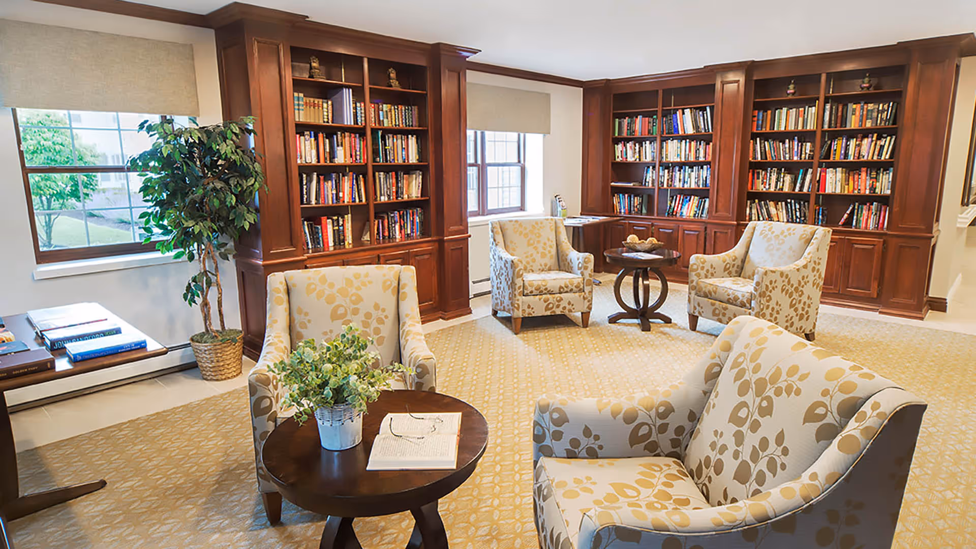A cozy senior living common area with floral patterned armchairs arranged around small wooden tables. The room features large wooden bookshelves filled with books, two windows with beige blinds, a potted plant, and a beige patterned carpet.