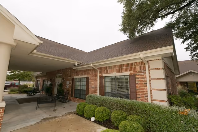 Exterior view of a brick building with a covered entrance, windows with shutters, and neatly trimmed bushes along the front. The sky is overcast and there are trees visible in the background.