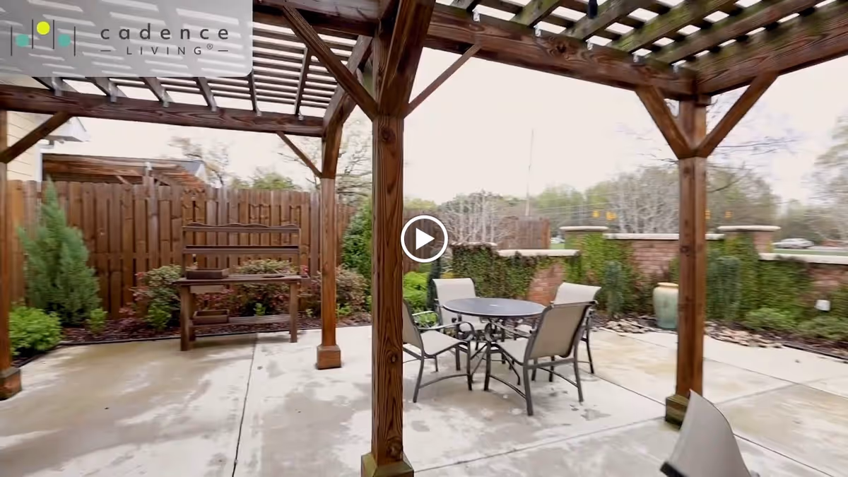 Outdoor patio area with a wooden pergola, a round table and chairs on a concrete slab and plants along a fenced perimeter.