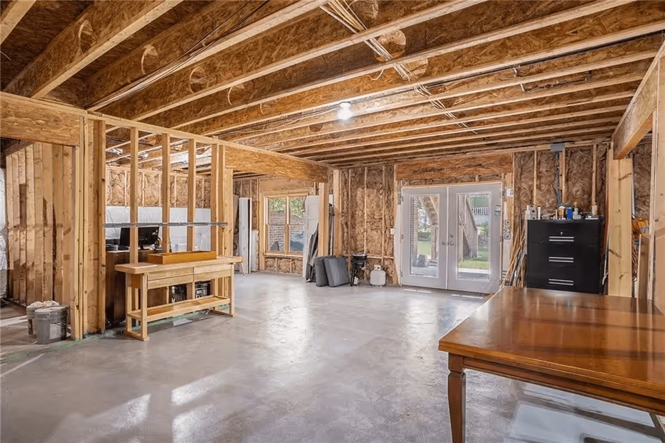 An unfinished interior room with exposed wooden beams and insulation on the walls. The floor is concrete, and there are double glass doors leading outside. Some furniture and construction materials are scattered around the space.