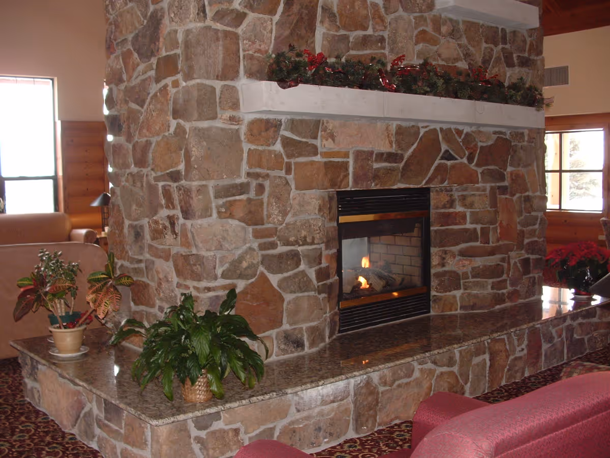 A cozy living room area featuring a large stone fireplace with a fire burning inside. The fireplace has a granite hearth with potted plants placed on it. There are windows in the background letting in natural light, and part of a red upholstered chair is visible in the foreground.