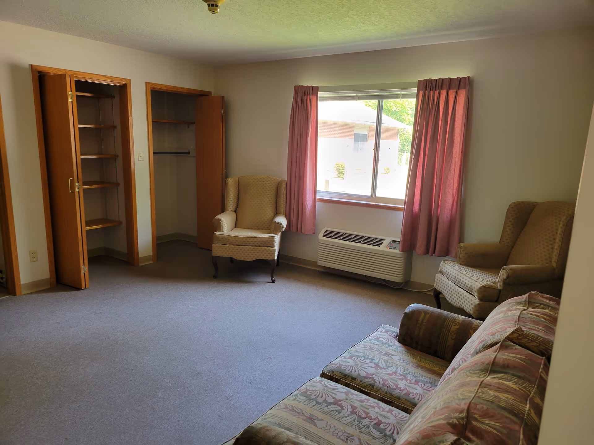 A small living room with two beige armchairs and a patterned sofa. There is a window with pink curtains letting in natural light, and below the window is a wall-mounted air conditioning unit. On the left side, there are two open closets with wooden doors and shelves inside. The room has light-colored walls and a gray carpeted floor.