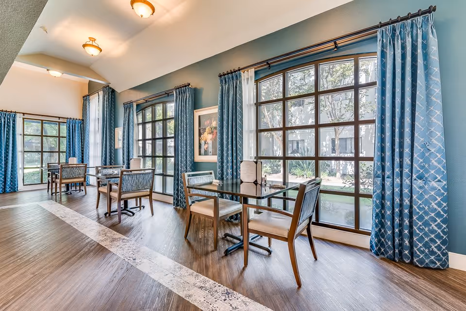 A bright dining area with large arched windows covered by blue patterned curtains, wooden tables with chairs, and a view of a garden outside. The room has a high ceiling with ceiling lights and a decorative tile strip on the wooden floor.