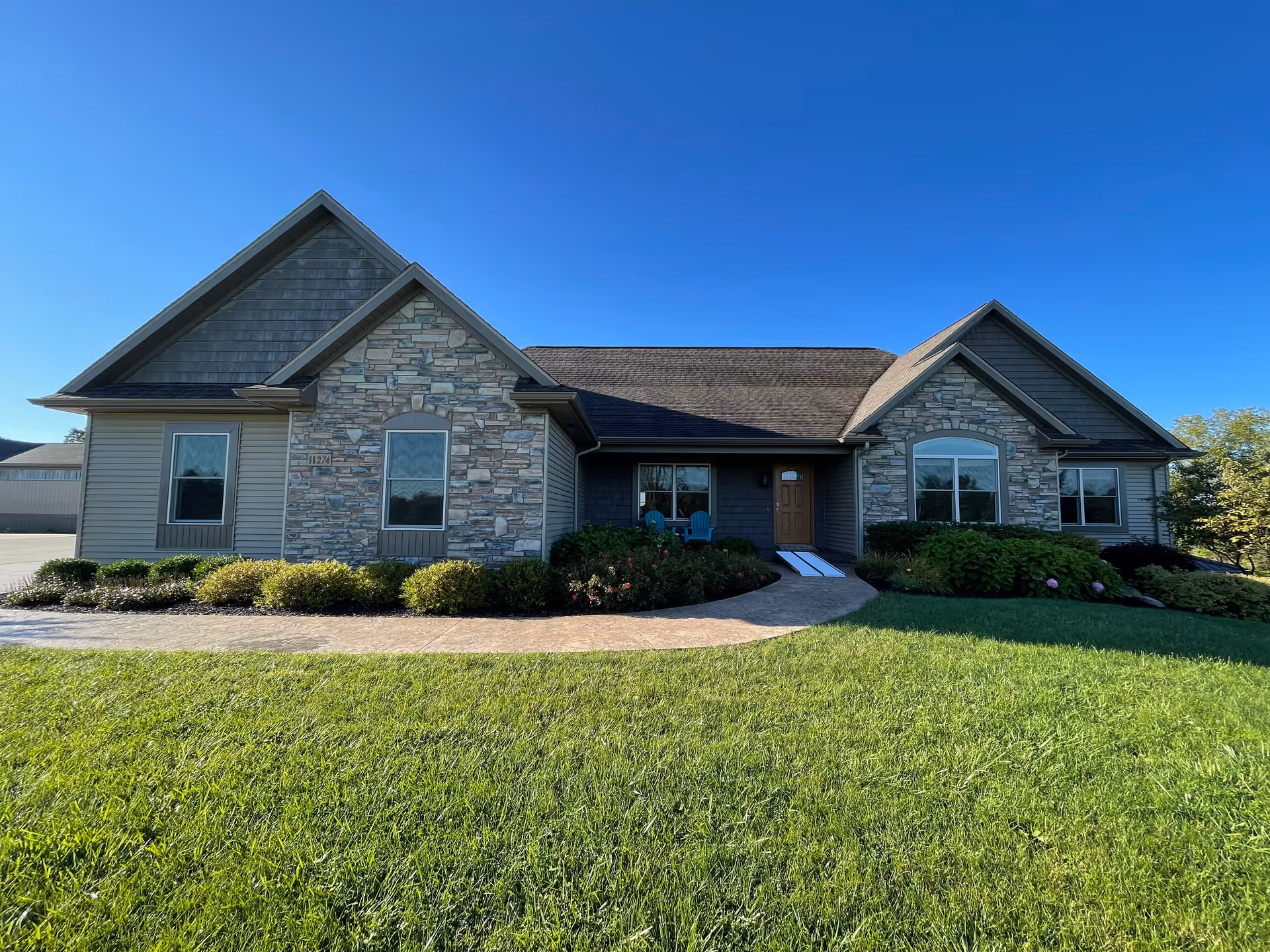 Front exterior of a single-story building with stone and siding, a walkway, lawn, and an entry with chairs.