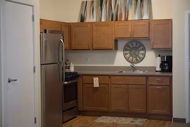 A kitchen area with wooden cabinets, a stainless steel refrigerator and oven, a sink with a faucet, a coffee maker, a paper towel holder, a wall clock with Roman numerals, and a colorful abstract painting above the cabinets.