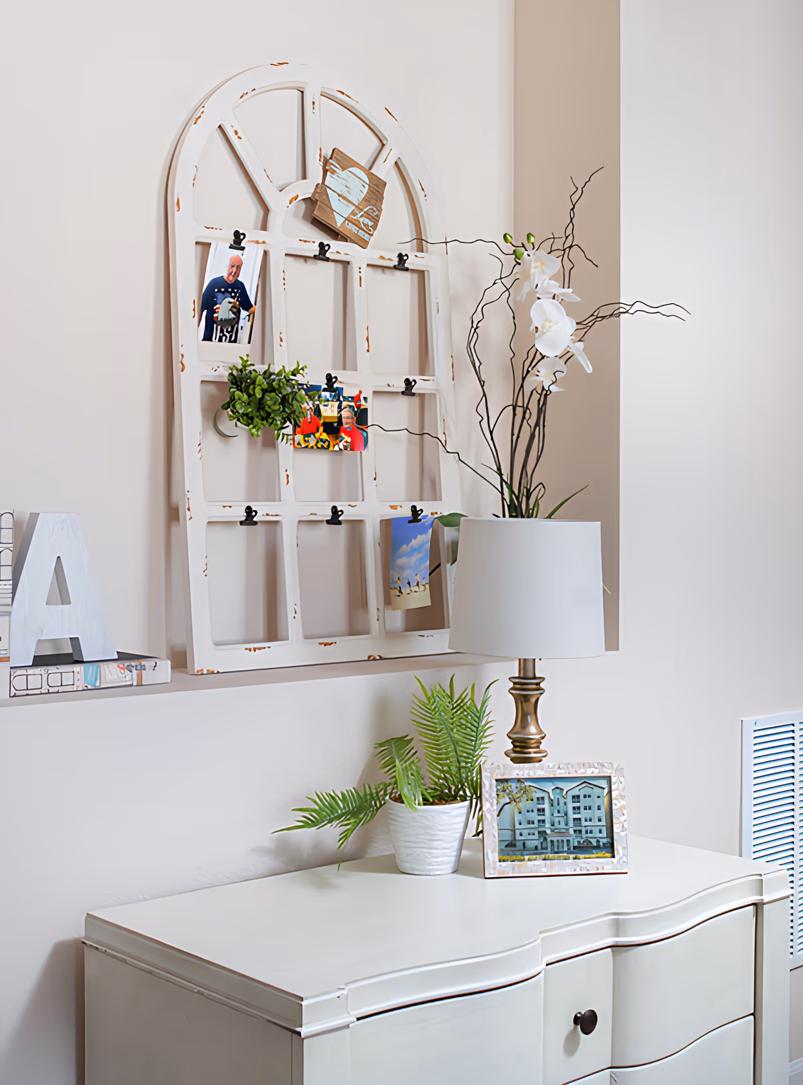 A white dresser with a small potted fern, a table lamp with a white shade, and a framed photo of a building on top. Above the dresser is a decorative white distressed window frame with clips holding photos and a small green plant. The wall behind is light beige.