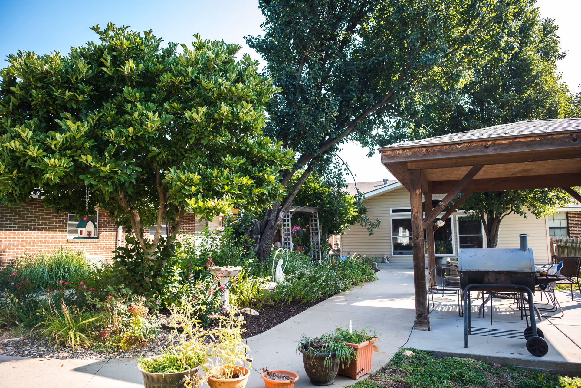 Outdoor garden area at Louisburg Healthcare and Rehabilitation Center featuring lush green trees, various plants and flowers, a birdhouse, a birdbath, and a covered patio with a barbecue grill and chairs.