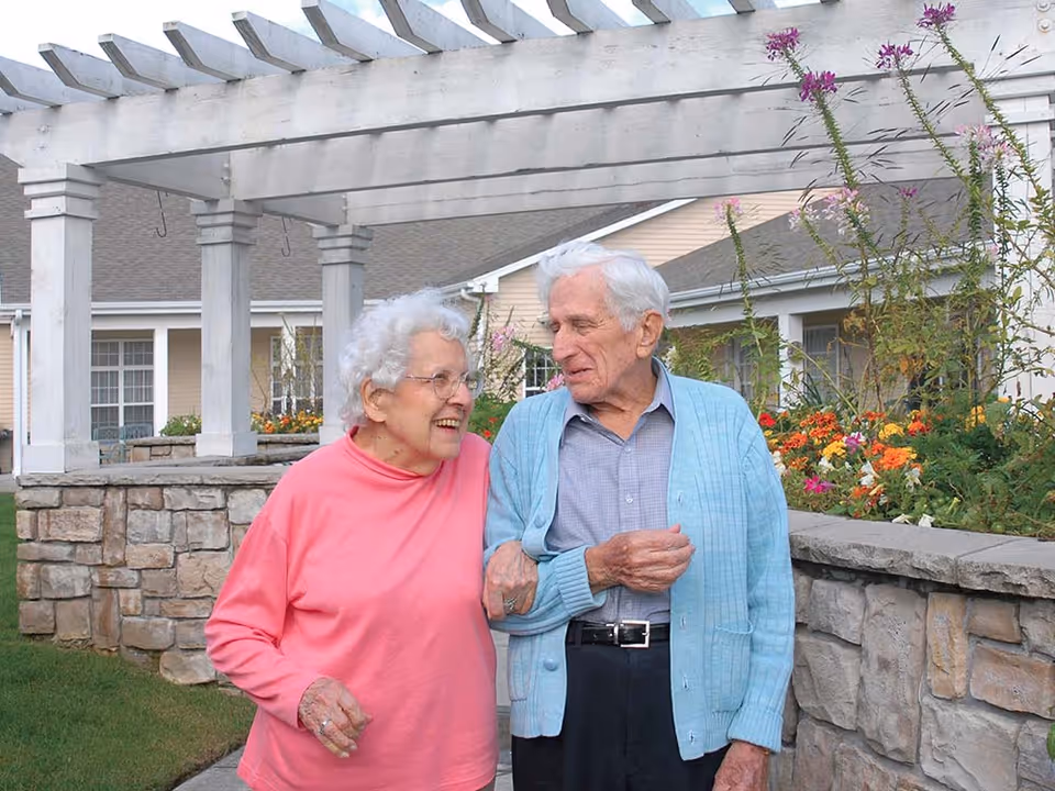An elderly woman in a pink sweater and an elderly man in a blue cardigan walk arm in arm outdoors near a stone wall and garden with flowers, under a white pergola structure with residential buildings in the background.
