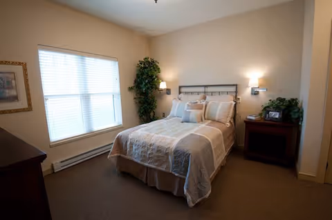 A cozy bedroom with a neatly made bed featuring a beige and white quilt and multiple pillows. The room has beige walls, a large window with blinds, a framed picture on the wall, a tall green plant in the corner, and a wooden nightstand with a lamp and some decorative items.