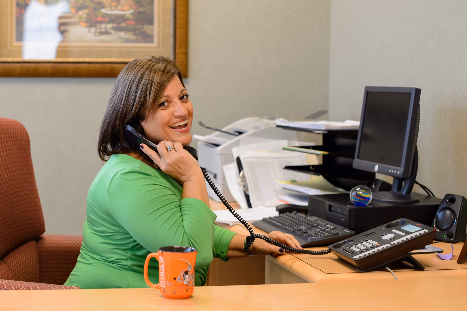 A woman in a green shirt sitting at a desk in an office, smiling while talking on a corded telephone. The desk has a computer monitor, keyboard, papers, a printer, and an orange Cleveland Browns coffee mug.