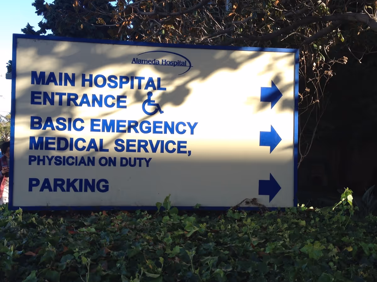 Outdoor sign at Alameda Hospital showing directions to the main hospital entrance, basic emergency medical service with physician on duty, and parking, with arrows pointing to the right. The sign is surrounded by greenery and partially shaded by tree branches.