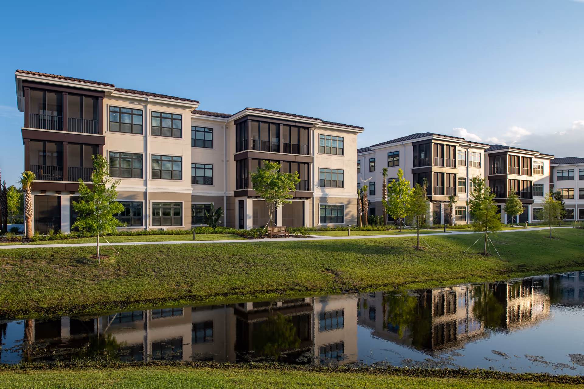Exterior view of The Mayflower at Winter Park senior living facility showing multiple three-story buildings with large windows and screened balconies, surrounded by green lawns, young trees, and a pond reflecting the buildings under a clear blue sky.