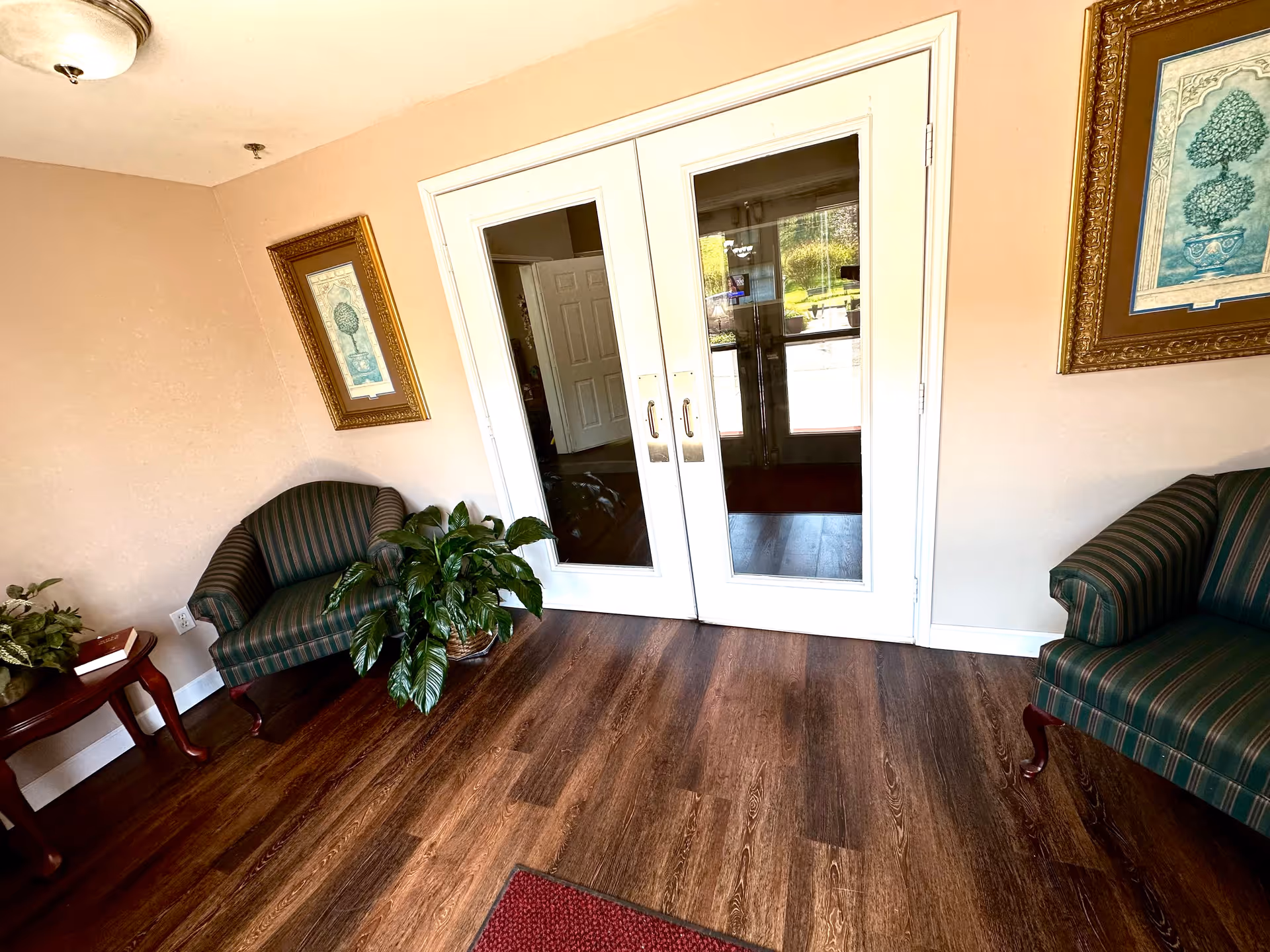 Interior view of a sitting area with two striped armchairs on either side of white double doors with glass panels. There are framed pictures on the beige walls and potted plants on the floor and a small wooden table with a book on it. The floor is wooden with a small red rug partially visible.