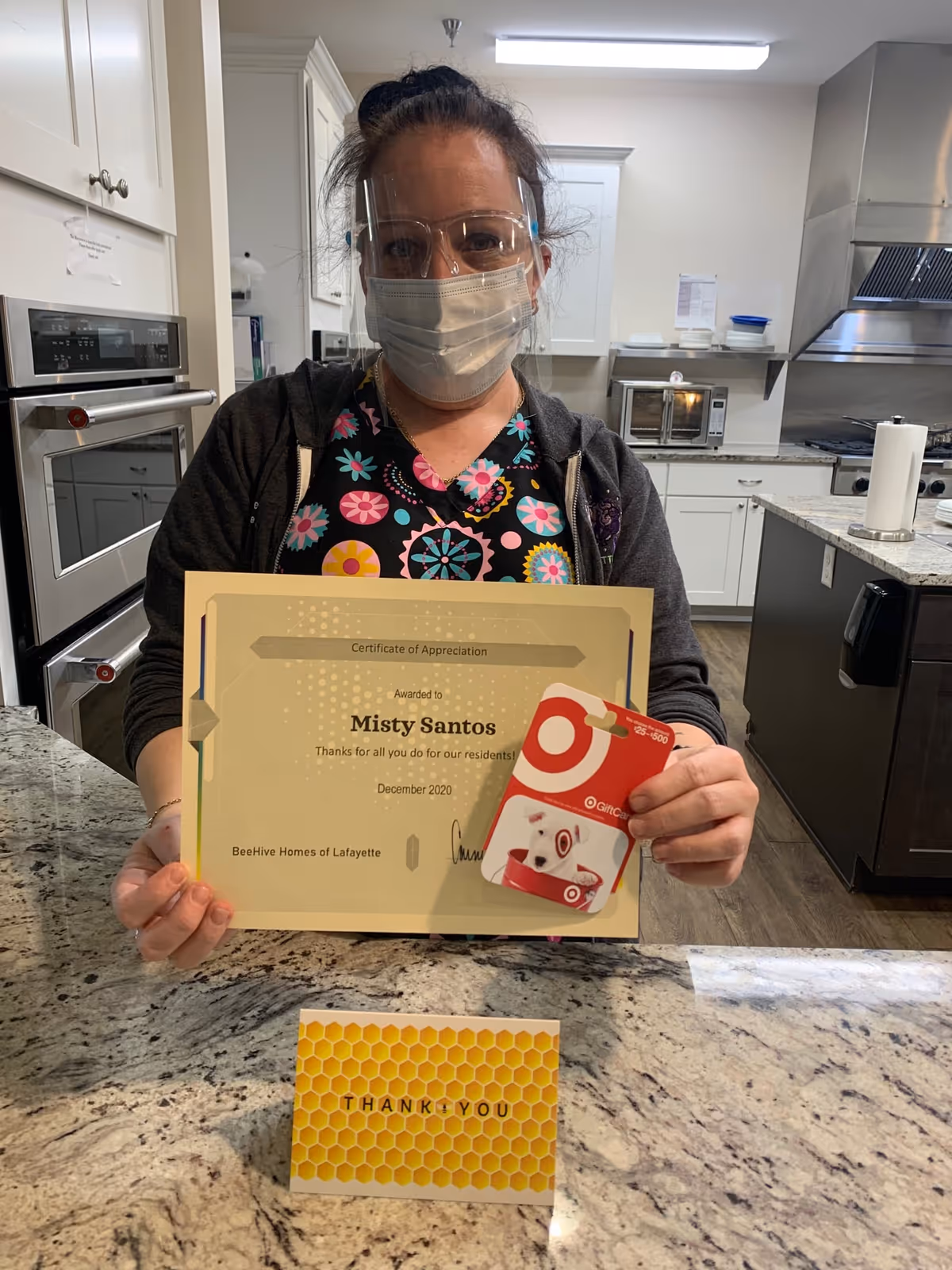 A woman wearing a face mask and protective glasses is standing in a kitchen holding a certificate of appreciation awarded to Misty Santos and a Target gift card. A thank you card with a honeycomb design is placed on the countertop in front of her.
