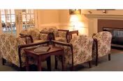 A cozy seating area with four floral upholstered armchairs arranged around a wooden coffee table with a book on it. A lit table lamp sits on a side table next to the chairs, and a fireplace is visible in the background. The room has a warm, inviting atmosphere.