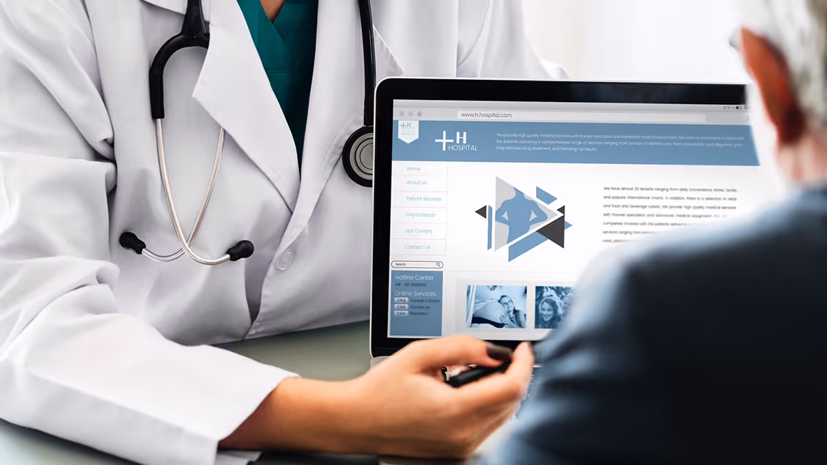 A medical professional wearing a white coat and stethoscope is showing information on a laptop screen to an elderly person. The laptop screen displays a hospital website with various sections and images.