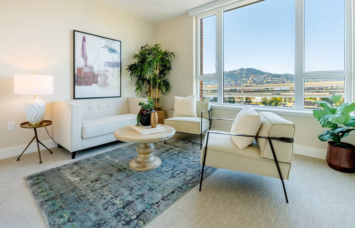 A bright living room with a large window showing a view of hills and a bridge outside. The room features a beige sofa, two beige armchairs with white cushions, a round wooden coffee table with decorative items, a side table with a white lamp, a large abstract painting on the wall, and two potted plants.