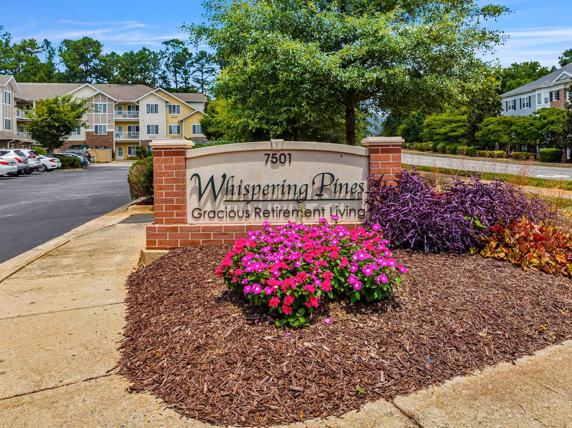 Entrance sign for Whispering Pines Gracious Retirement Living surrounded by landscaped flowers and apartment buildings in the background.