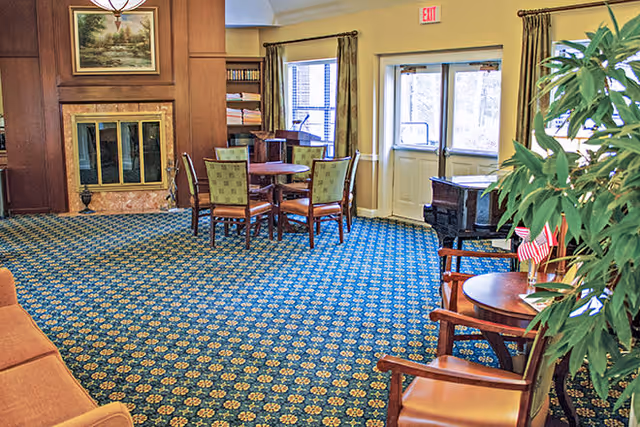 A cozy living room area in a senior living facility featuring a patterned blue carpet, a fireplace with a framed painting above it, a round table with four chairs, a bookshelf, a piano near the exit doors, and several chairs around small tables with plants and American flags.