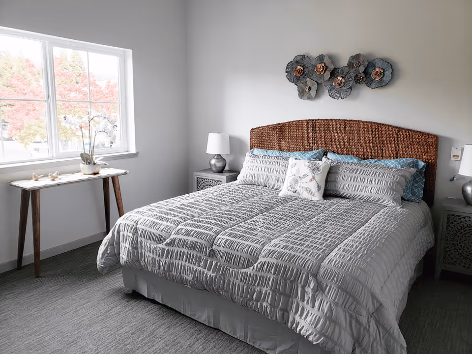 A neatly made bed with a textured gray comforter and multiple pillows in a bright bedroom. The bed has a woven headboard and is flanked by two matching nightstands with silver lamps. A decorative metal wall art piece hangs above the headboard. A wooden table with a potted orchid sits under a large window showing colorful trees outside.