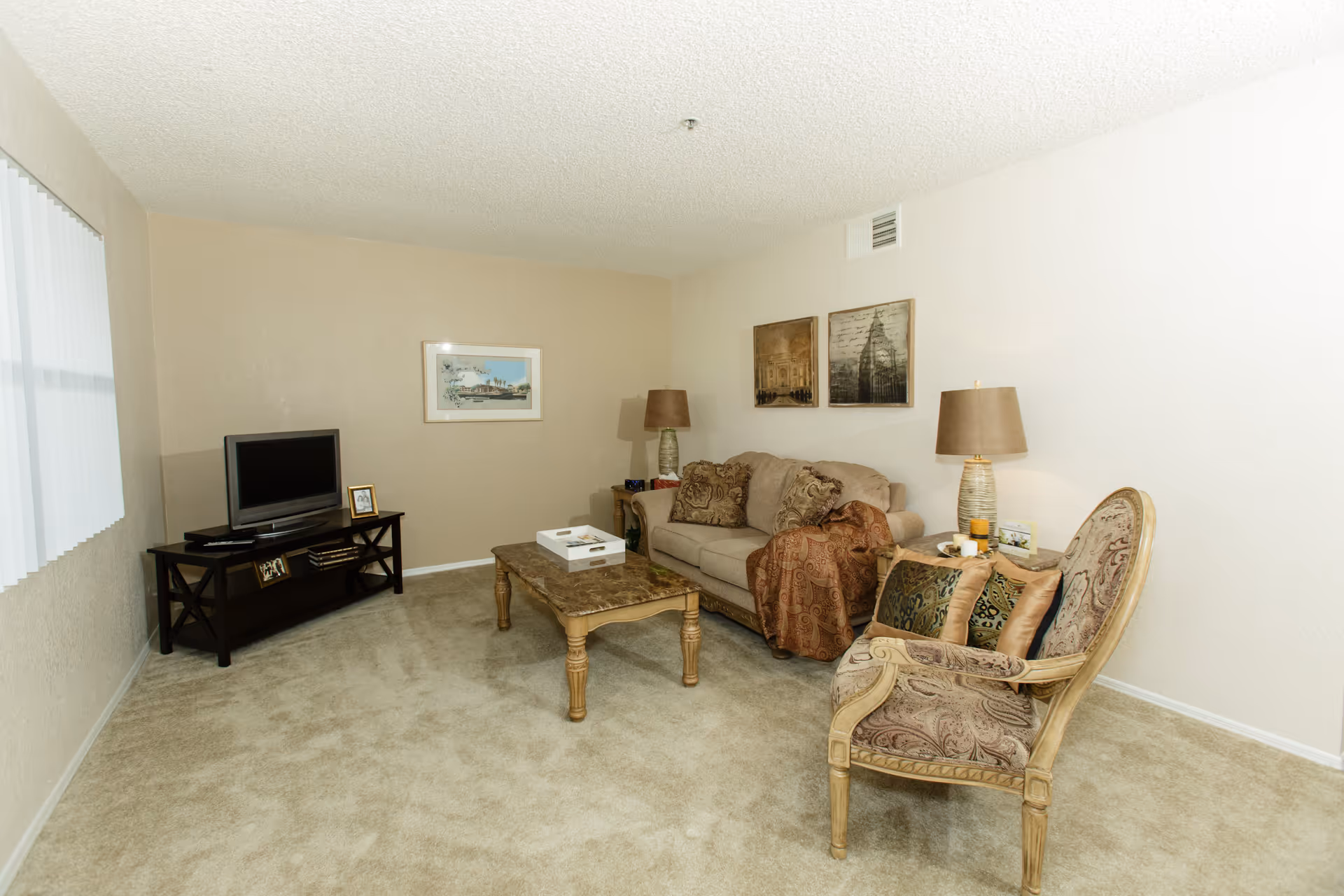 A cozy living room with beige walls and carpet. The room features a beige sofa with patterned cushions and a throw blanket, a wooden coffee table with a decorative tray, an upholstered armchair with two pillows, two table lamps on side tables, a TV on a dark wooden stand, and framed artwork on the walls.